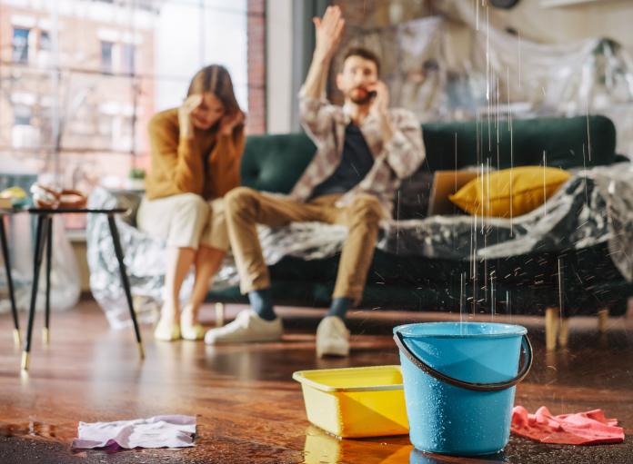 A man and woman sit on a couch in their living room with water dripping from the ceiling into buckets; the man is on the phone with a plumber in Denville NJ while the woman looks stressed.