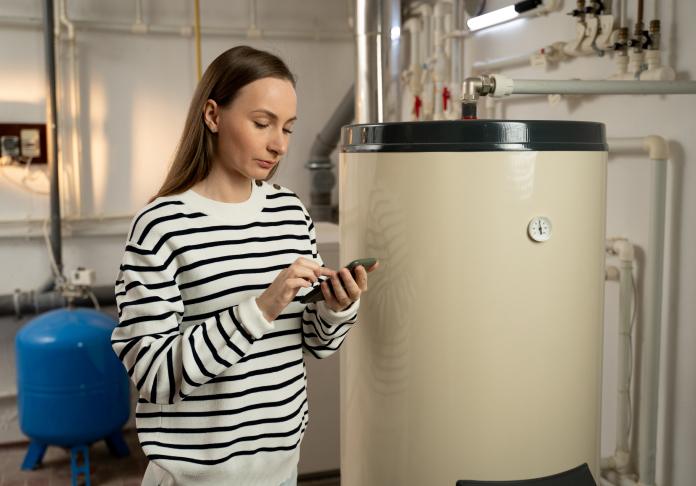 A woman in a striped sweater uses her smartphone while standing next to a large water heater—perhaps searching for plumbing in West Milford NJ—in a utility room with pipes and a blue pressure tank in the background.