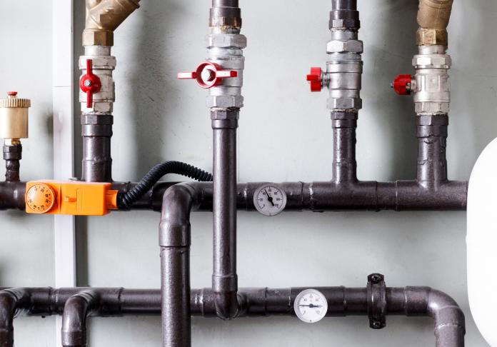 A set of black metal pipes with red valve handles, pressure gauges, and an orange electrical component is mounted on a light-colored wall—ideal for plumbing in Denville NJ, this network serves heating or plumbing needs efficiently.