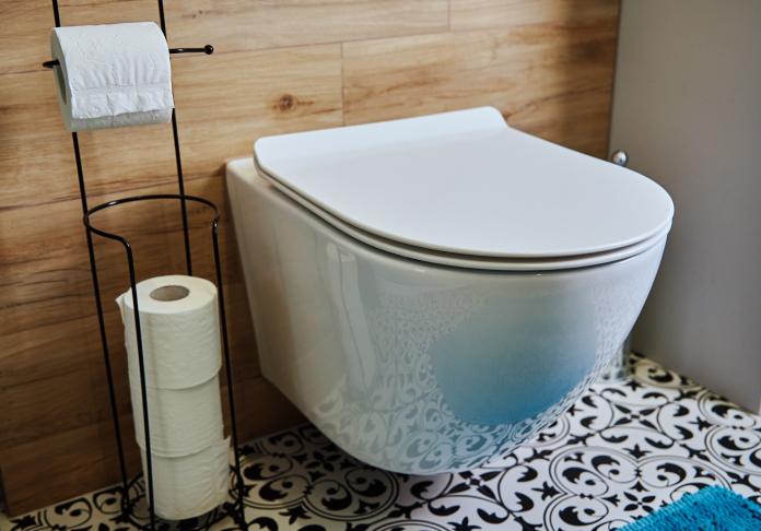 A modern white wall-mounted toilet with a closed lid, beside a black metal stand holding toilet paper, set against wood paneling and patterned tiles—perfect inspiration from a top plumber in Wayne NJ.