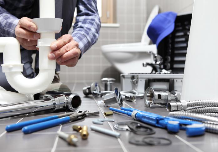 A plumber in Denville NJ installs white PVC pipes under a bathroom sink. Various plumbing tools and metal fittings are scattered on the tiled floor, with a toilet and toolbox visible in the background.