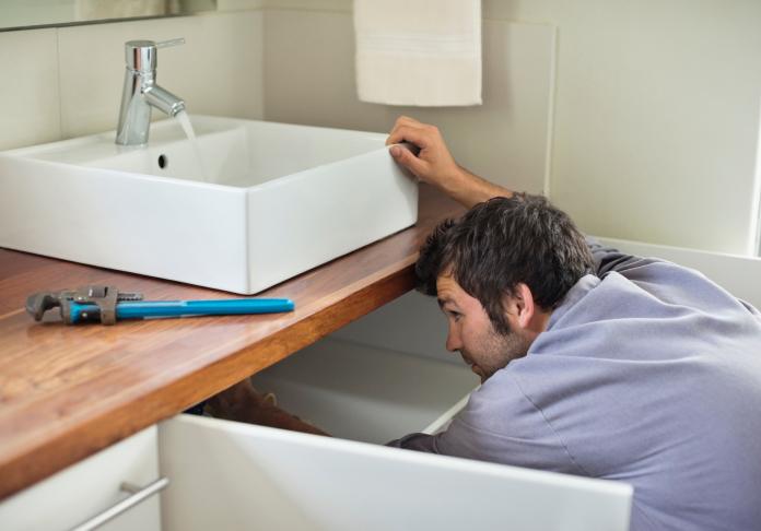 A man is crouched under a bathroom sink, inspecting the plumbing in West Milford NJ. A wrench lies on the wooden countertop next to the white ceramic sink, and the cabinet drawers are open as he works.
