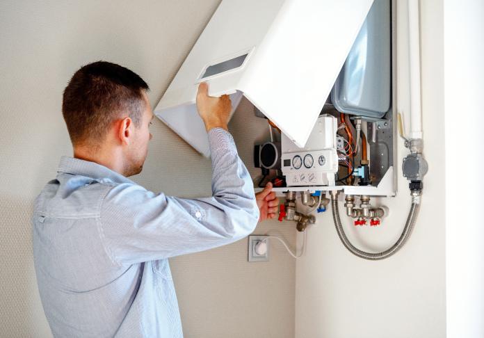A man, possibly a plumber in Wayne NJ, adjusts or repairs a wall-mounted gas boiler, lifting its front panel to access internal components and pipes in a home utility area.