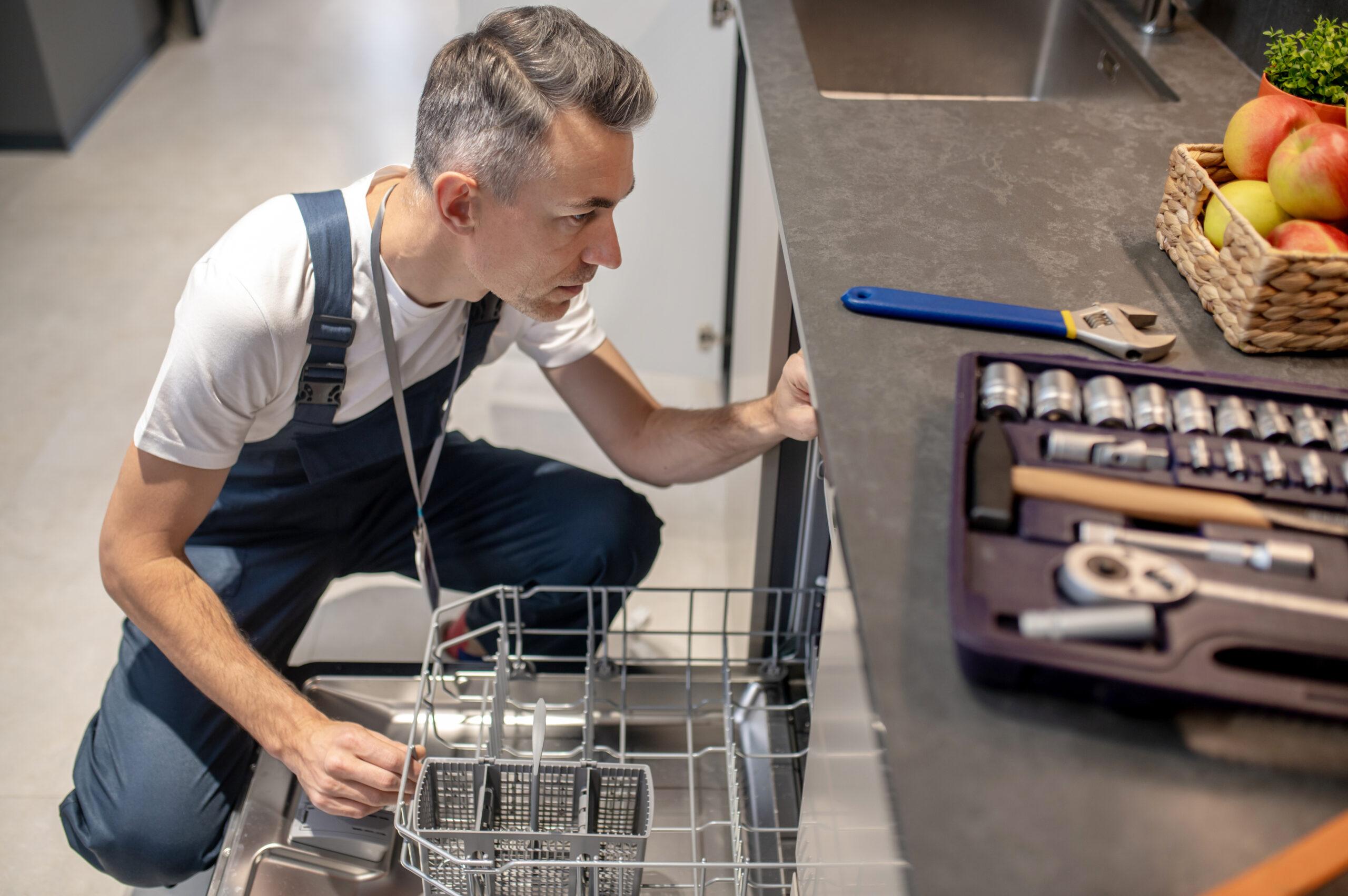 A repairman in overalls inspects an open dishwasher in a kitchen. Tools, including a wrench and a toolkit, are laid out on the countertop nearby. A basket of fruit is also visible on the counter.
