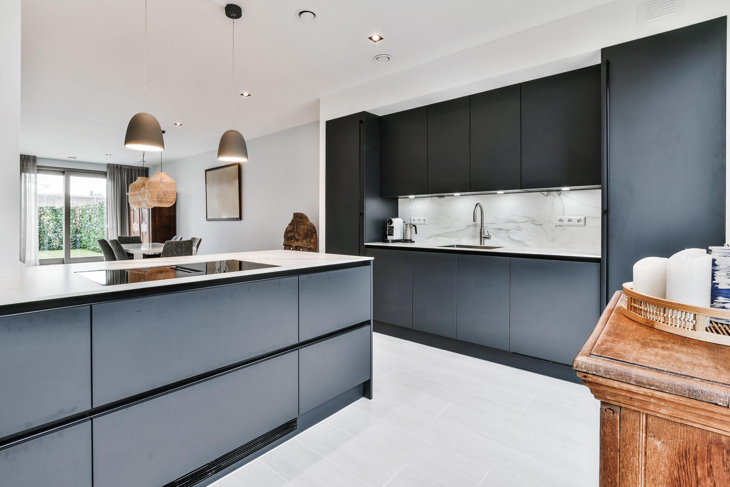 Modern kitchen with sleek black cabinets, white countertops, built-in appliances, and pendant lighting. A dining area with a table and chairs is visible in the background near large windows with curtains.