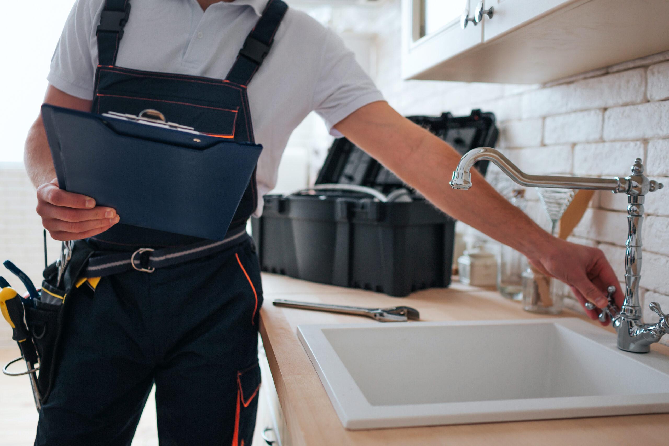 A plumber in overalls holds a clipboard and adjusts a kitchen sink faucet, with a toolbox and tools on the countertop.