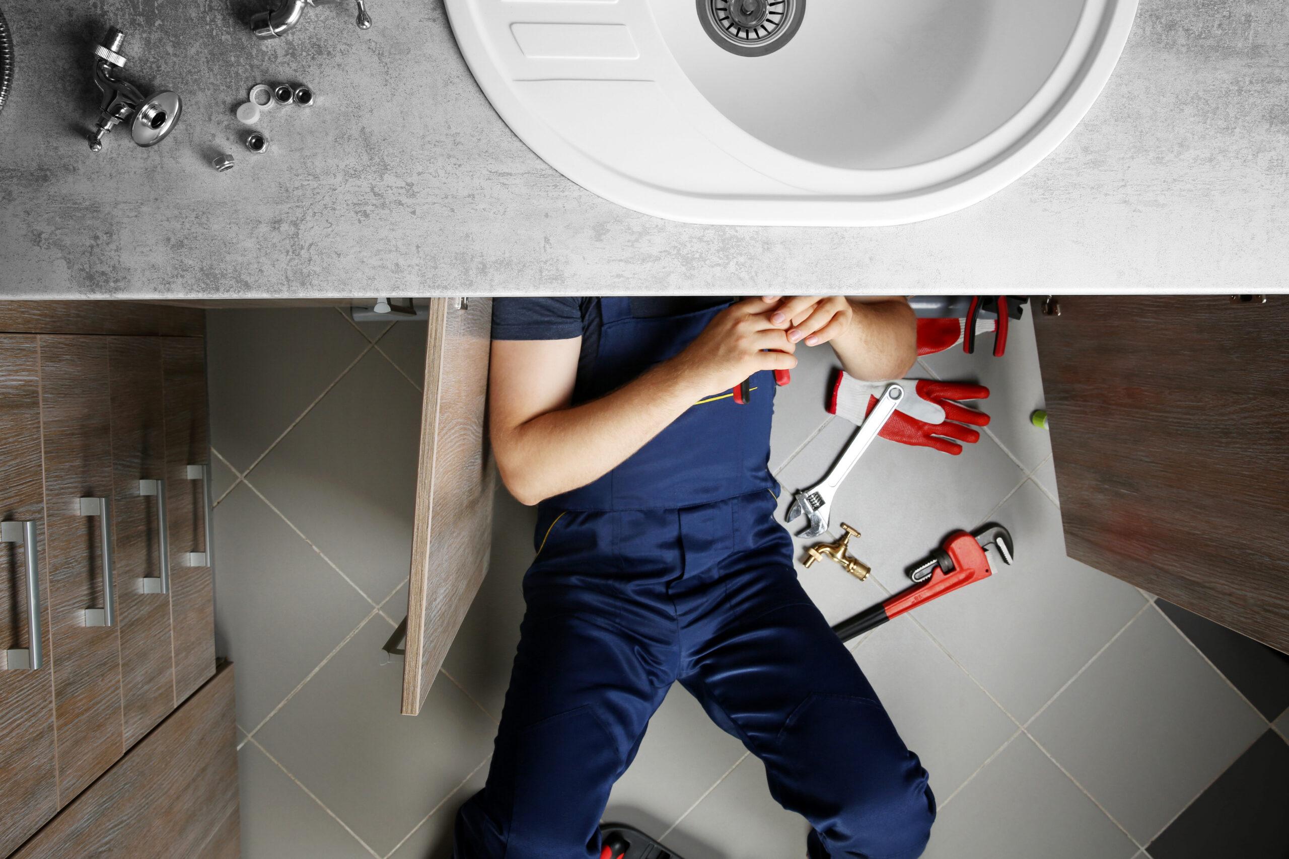 A plumber in blue overalls lies on the floor under a kitchen sink, working on the pipes. Tools and plumbing parts are scattered around him on the tiled floor.