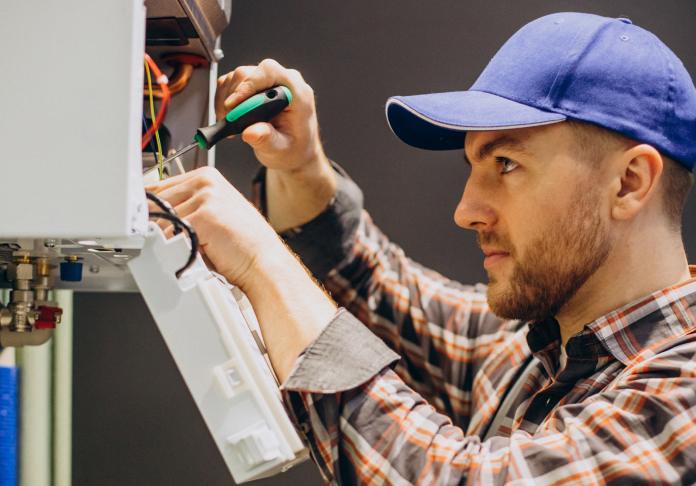 A man wearing a blue cap and plaid shirt uses a screwdriver to repair or install an electrical appliance, focusing intently on the wiring inside an open panel.