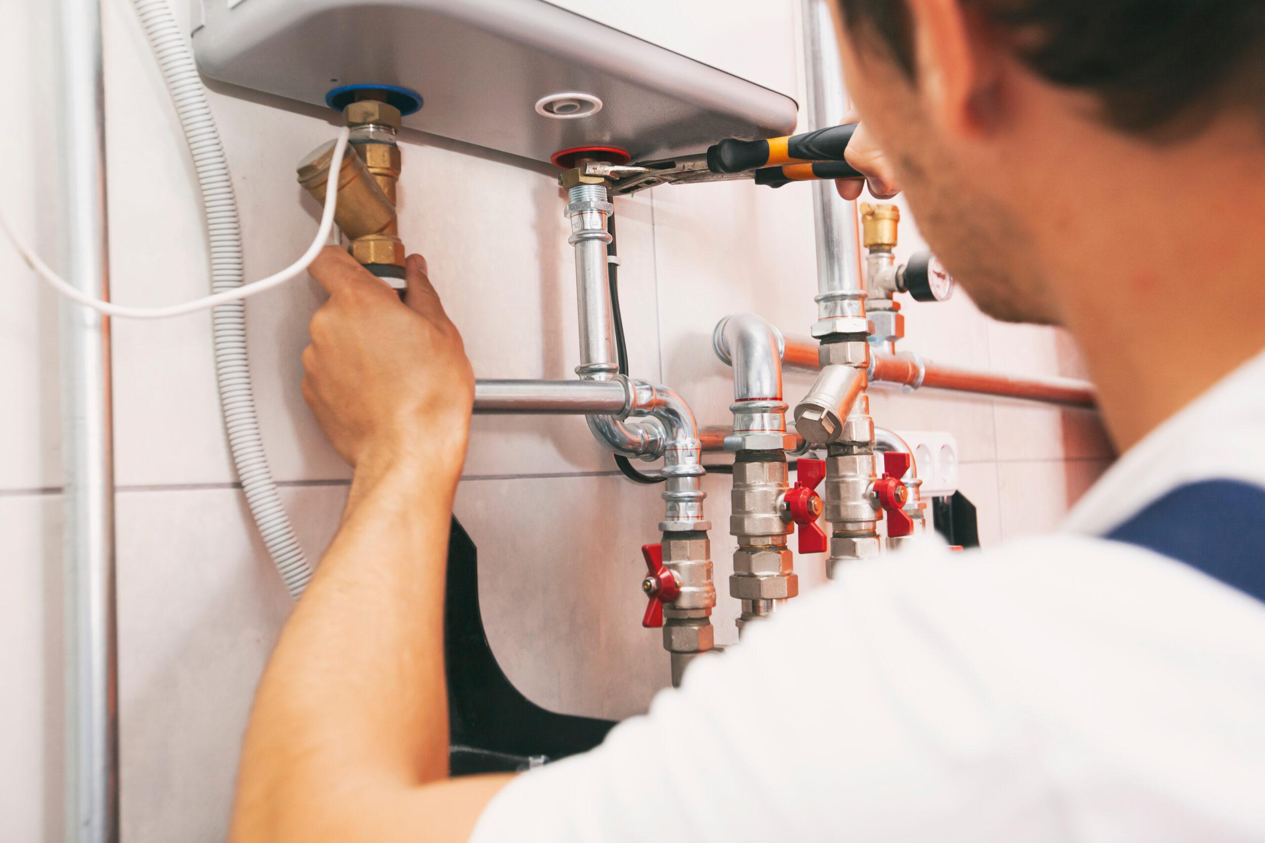A plumber wearing a white shirt adjusts a valve under a sink, surrounded by pipes, fittings, and plumbing tools on a tiled wall.