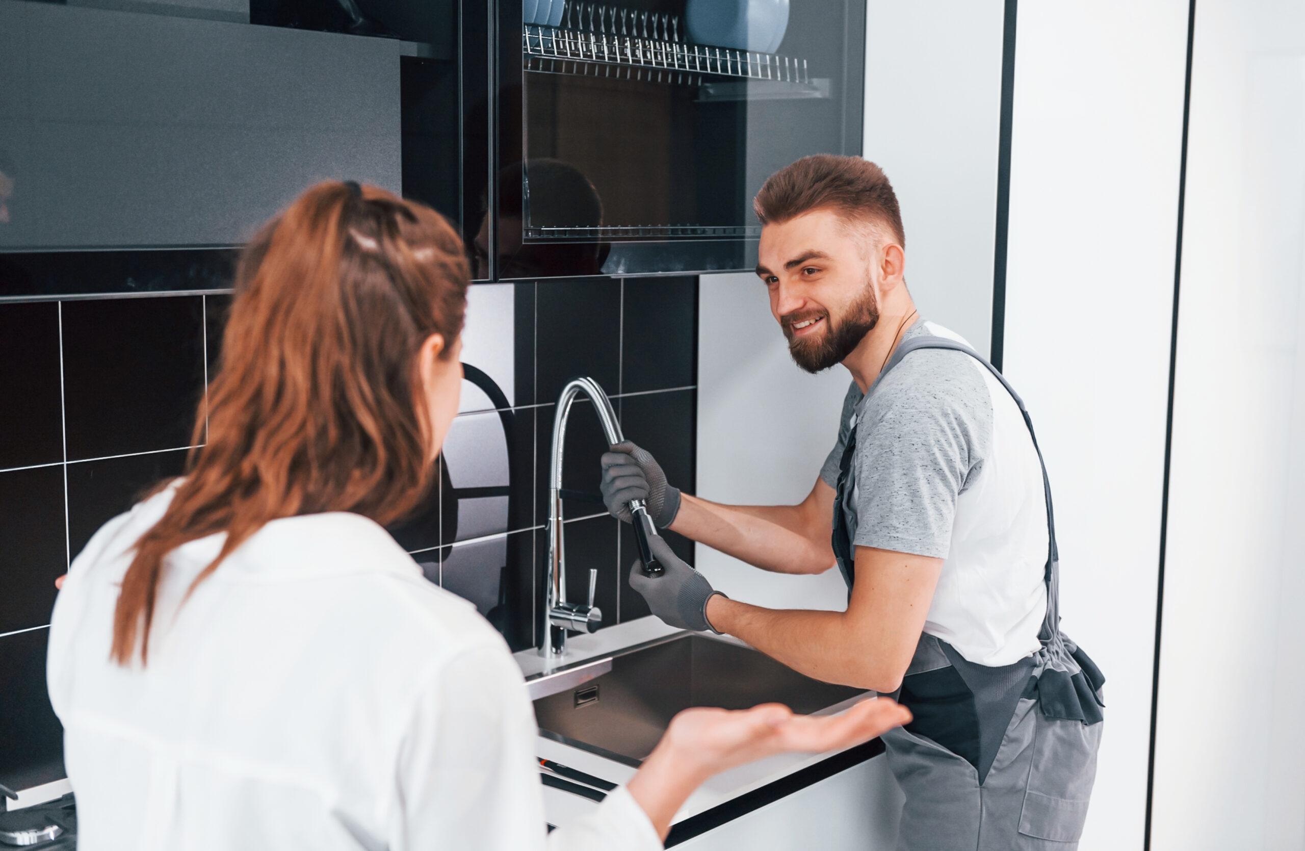 A plumber in work clothes and gloves is fixing a kitchen sink faucet while smiling at a woman who is talking to him. The modern kitchen features black tiles and white cabinets.
