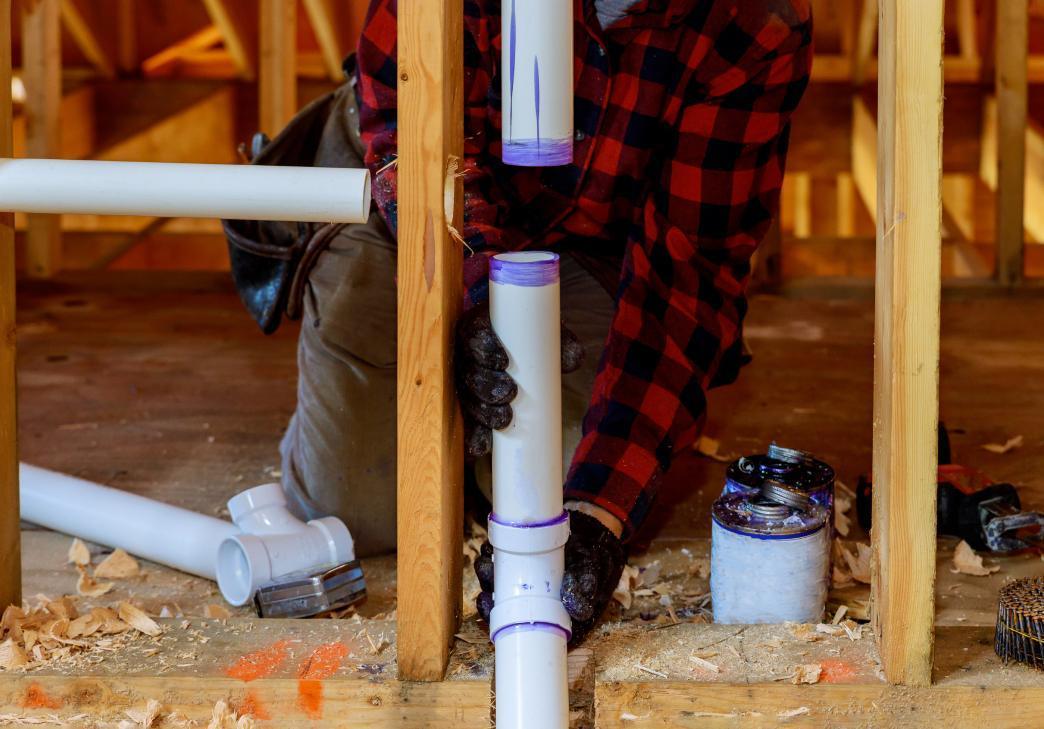 A person wearing gloves and a red plaid shirt assembles white PVC pipes in a wooden-framed building under construction, with tools and pipe fittings nearby.