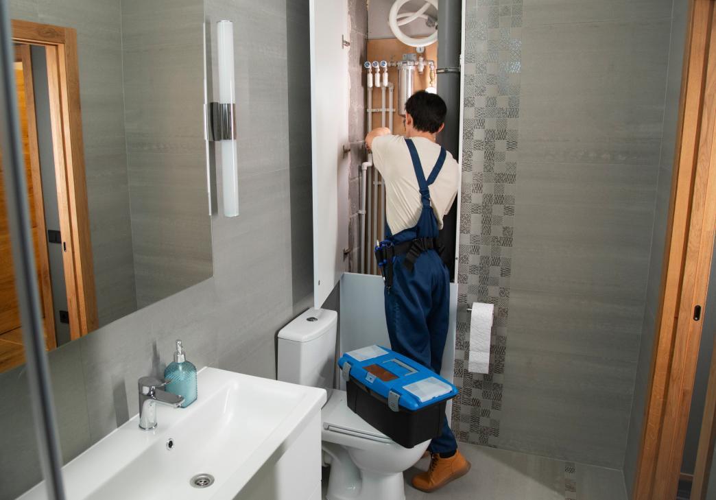 A plumber in overalls works on pipes inside a bathroom, standing near a toilet and sink. A blue toolbox is placed on the closed toilet lid. The bathroom has gray walls and modern fixtures.