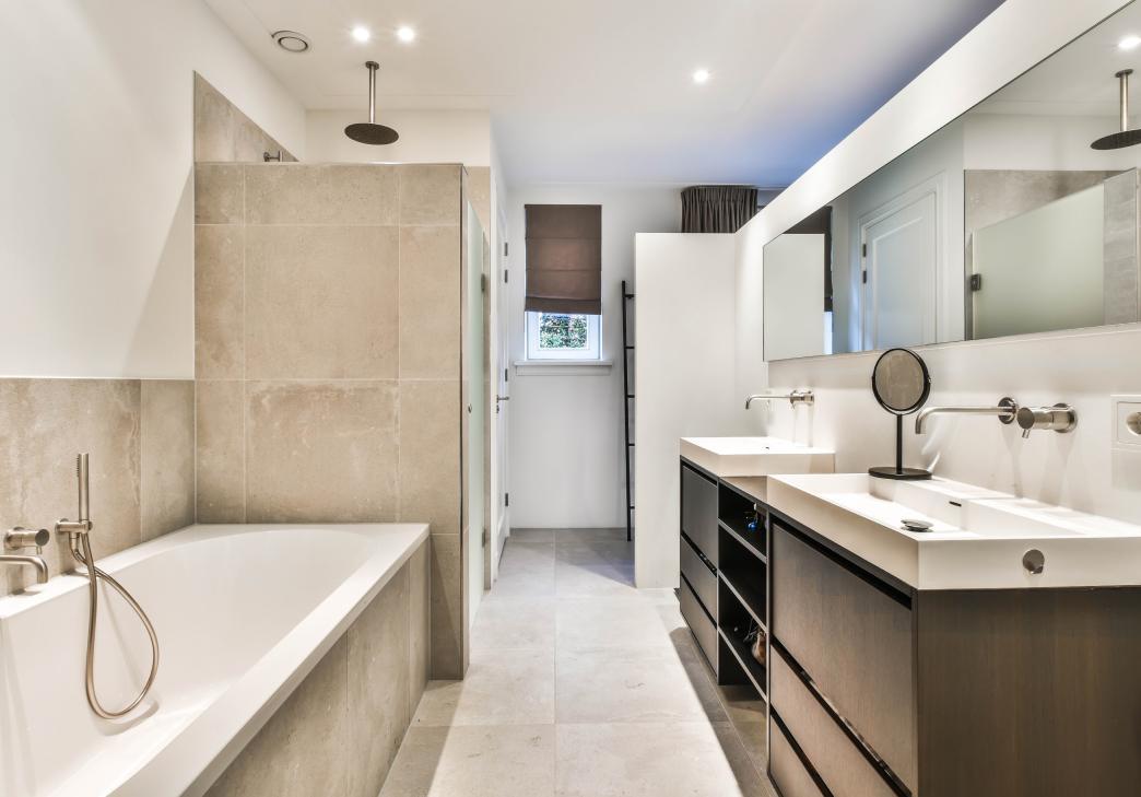 Modern bathroom with beige tiles, a white bathtub on the left, a glass-enclosed shower, double sinks with large mirrors, dark wood cabinets, and a small window with a brown blind.