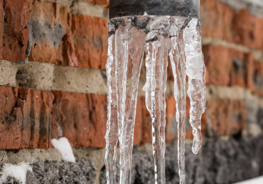 Several clear icicles hang from the end of a metal pipe against a background of a brick wall with patches of snow.