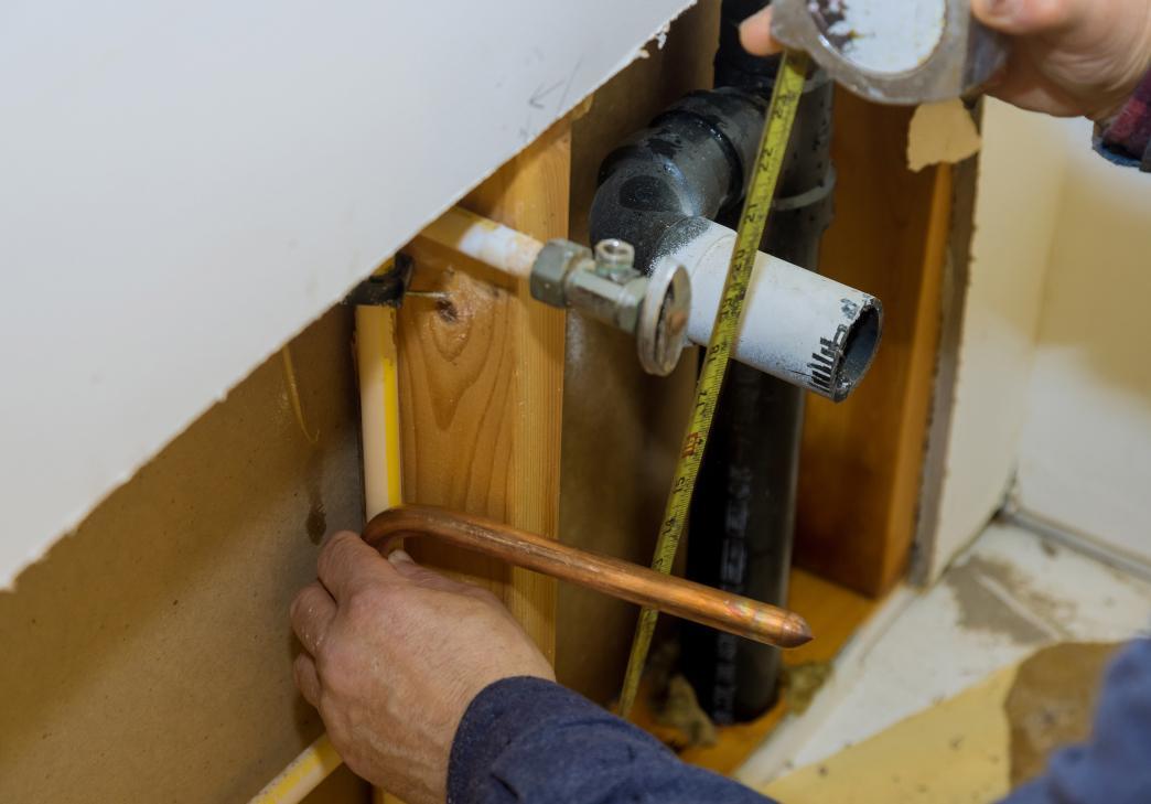 A person measures pipes inside a wall during plumbing repairs, using a measuring tape and holding a copper pipe near exposed wooden studs and various plumbing lines.