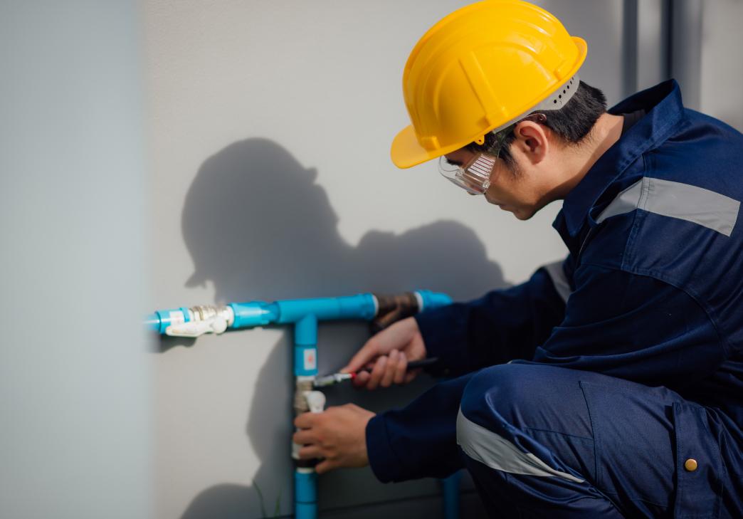 A worker wearing a yellow safety helmet and blue coveralls uses a tool to adjust blue water pipes attached to a wall.