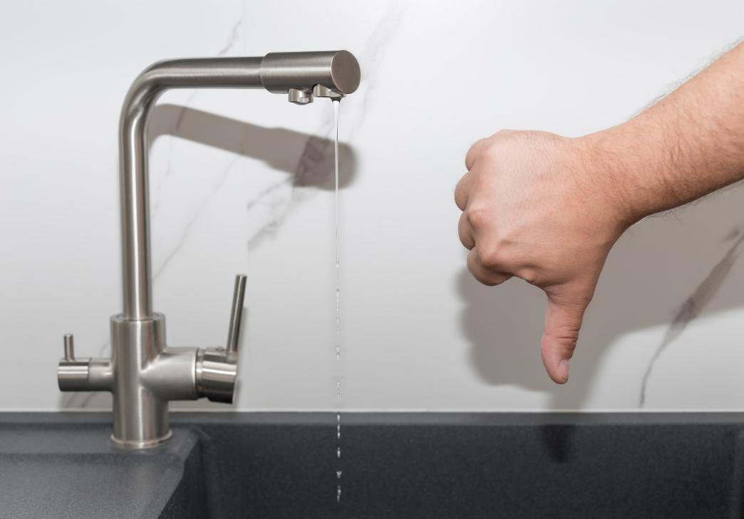 A person gives a thumbs-down gesture next to a kitchen faucet with a very weak stream of water, suggesting dissatisfaction with low water pressure.
