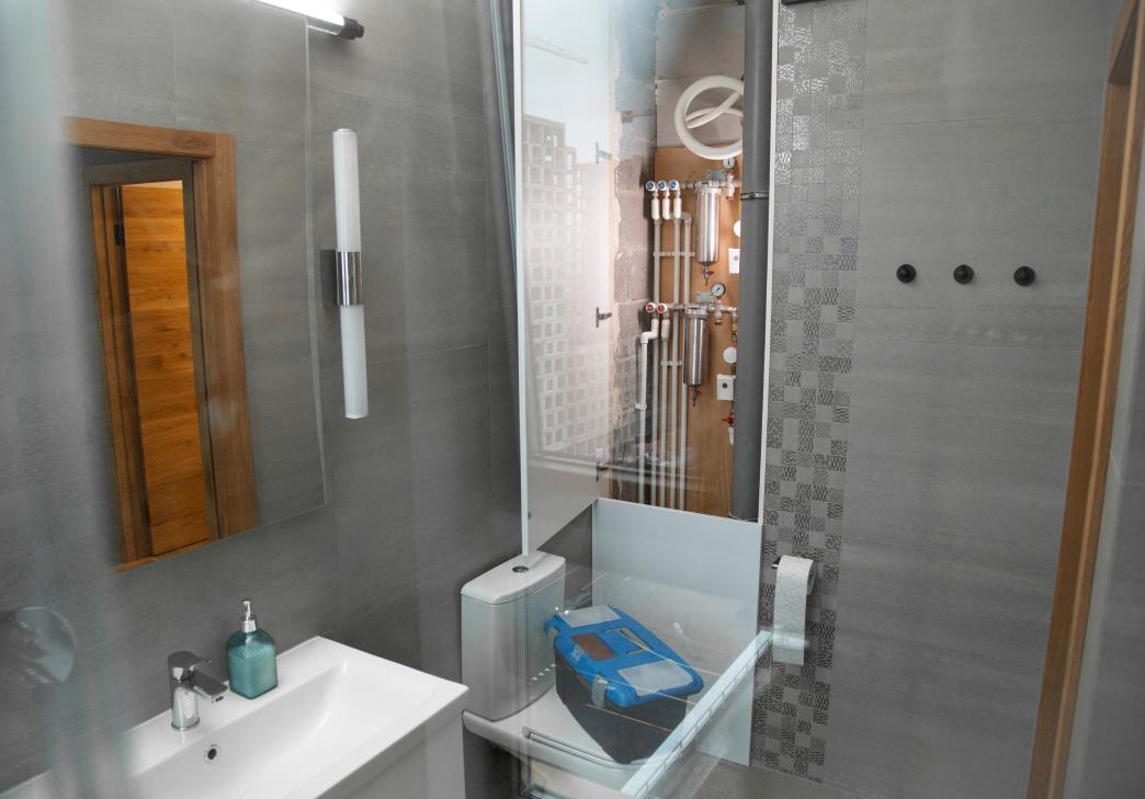 Modern bathroom with gray tiles, a white sink, and a mirror. A toilet is next to a bathtub with a transparent screen. Plumbing pipes and meters are visible behind glass at the end of the bathtub. Soap and sanitizer are on the sink.