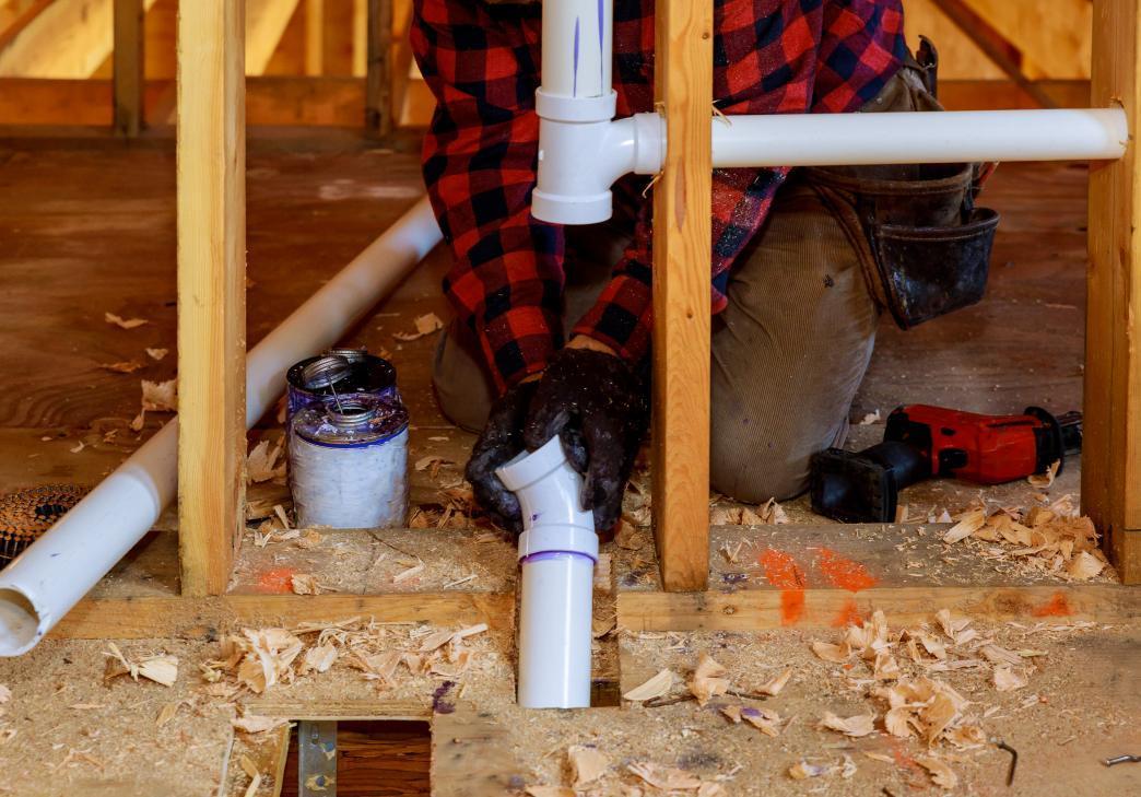 A person wearing a red plaid shirt and gloves is installing white PVC pipes for plumbing in a wooden-framed building under construction. Tools, pipe glue, and wood shavings are scattered on the floor.