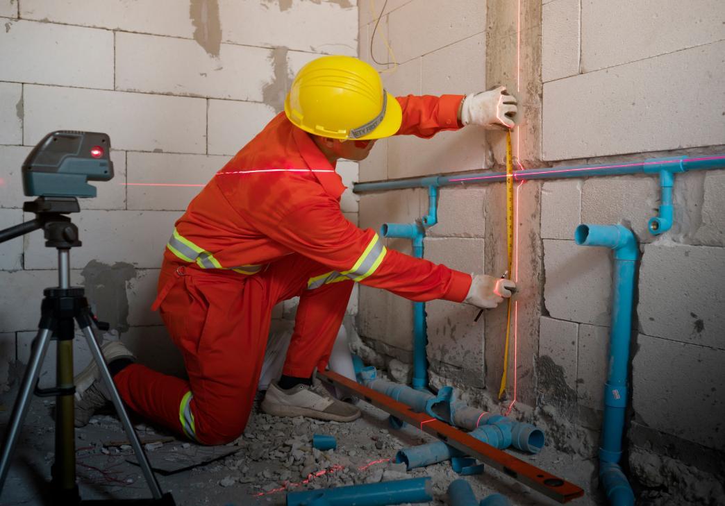 A construction worker in an orange safety suit and yellow helmet uses a laser level to align pipes on a wall, surrounded by blue PVC pipes in an unfinished room.