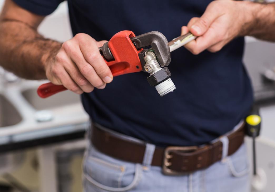 A person using a red pipe wrench to tighten a metal pipe fitting, wearing a dark blue shirt and jeans, with tools visible on their belt in the background.