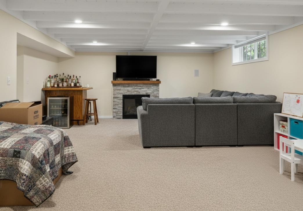 A finished basement with beige walls, a gray sectional sofa facing a TV on a stone fireplace, a bar area with stools and bottles, a bed with a quilt, and a children's table with toys. Light carpet and a small window complete the space.