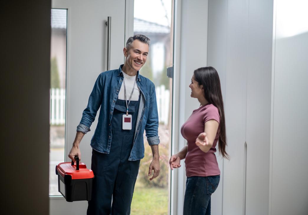 A smiling repairman with a toolbox enters a home while a woman in a pink shirt gestures toward the interior, welcoming him in through the open front door.
