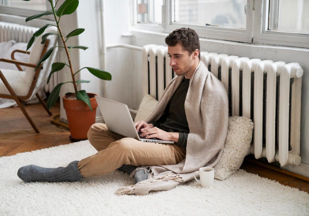 A man sits on a carpeted floor, wrapped in a blanket with a mug nearby, using a laptop in a bright room with large windows and a potted plant.