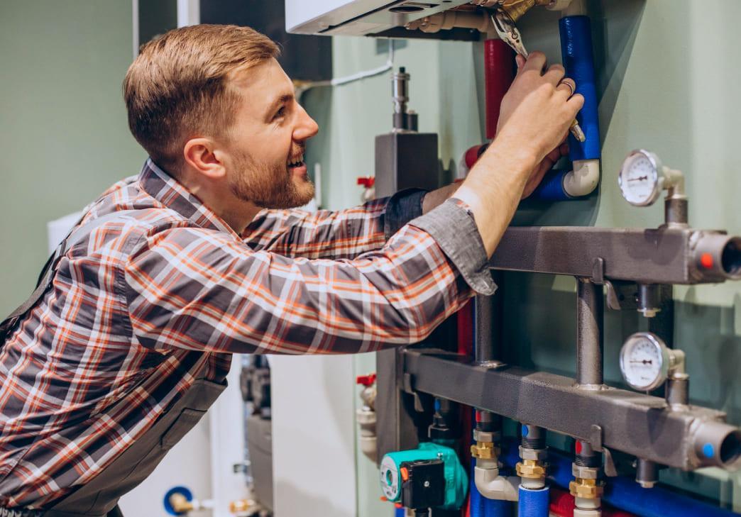 A smiling man in a plaid shirt adjusts pipes and valves on a heating or plumbing system mounted to a wall, with various gauges and pipes visible.