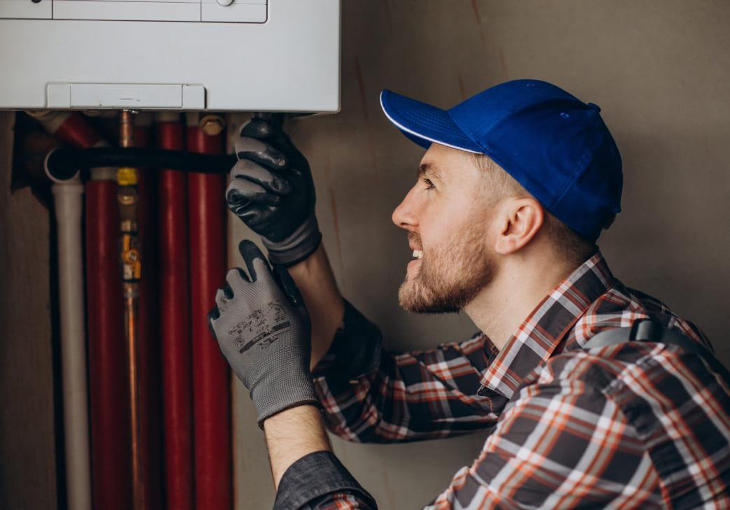 A smiling man wearing a blue cap, plaid shirt, and gloves is adjusting or repairing a wall-mounted boiler with red and black pipes visible in the background.