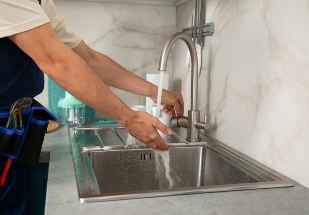 A person tests the water flow from a modern kitchen faucet over a stainless steel sink, with tools visible in their belt and a marble-patterned backsplash in the background.