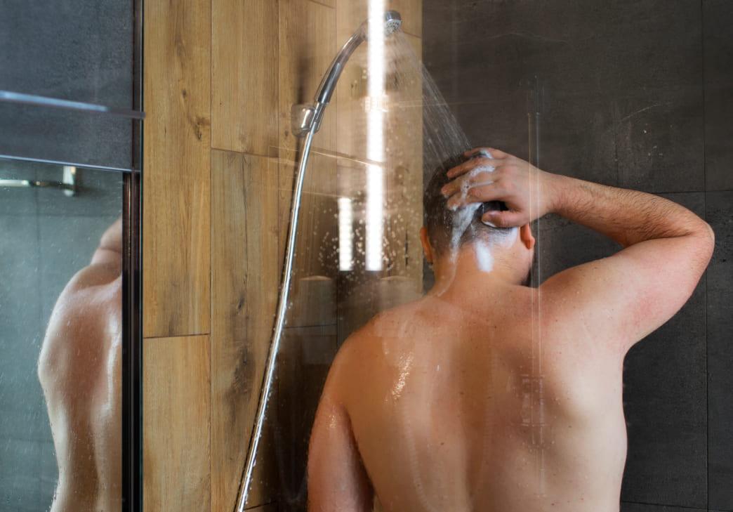 A person is standing in a shower with their back to the camera, washing their hair under running water. The shower has tiled walls in wood and dark gray tones.