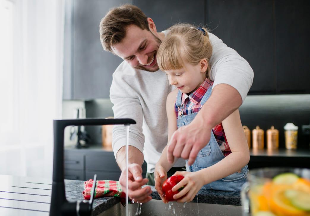 A man and a young girl smile as they wash a red bell pepper together under a kitchen faucet. The man is helping the girl, and they both appear happy, working in a modern kitchen.