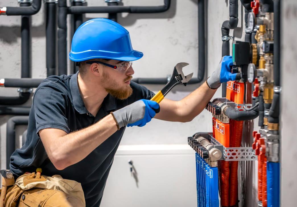 A plumber in a blue hard hat and gloves uses a wrench to adjust pipes and valves on a wall-mounted plumbing system. Various colored pipes and gauges are visible in an industrial setting.