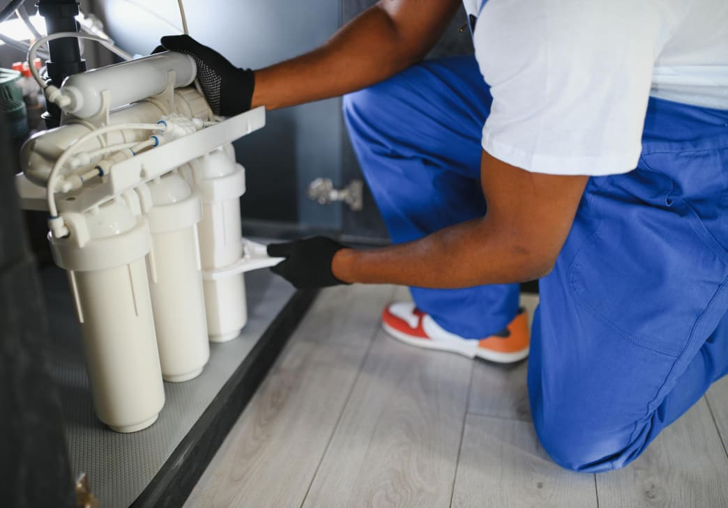 A person in blue overalls and gloves is kneeling on a wooden floor, installing or inspecting a water filtration system under a sink.