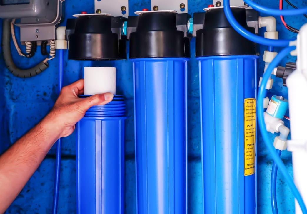 A person’s hand is installing or replacing a filter cartridge in a blue water filtration system with three cylindrical housings mounted on a wall.